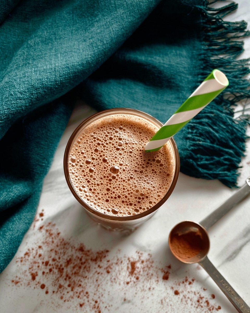 A tall transparent glass filled with a smooth, light brown smoothie that has small darker specks throughout. A green and white striped paper straw is placed inside the glass, standing upright. The glass is set on a white marbled surface lightly sprinkled with a fine brown powder around it. In the blurred background, a teal cloth is casually spread out. The photo taken with an iphone --ar 4:5 --v 7