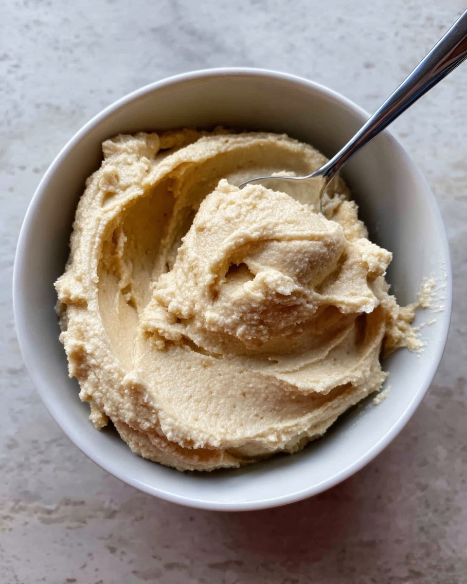 A white bowl filled with a creamy, thick, light beige spread that has a slightly grainy texture. The spread is heaped unevenly inside the bowl, showing soft peaks and small air pockets. A silver spoon is partially submerged on the right side, with some of the spread clinging to it. The bowl is placed on a white marbled texture surface. photo taken with an iphone --ar 4:5 --v 7
