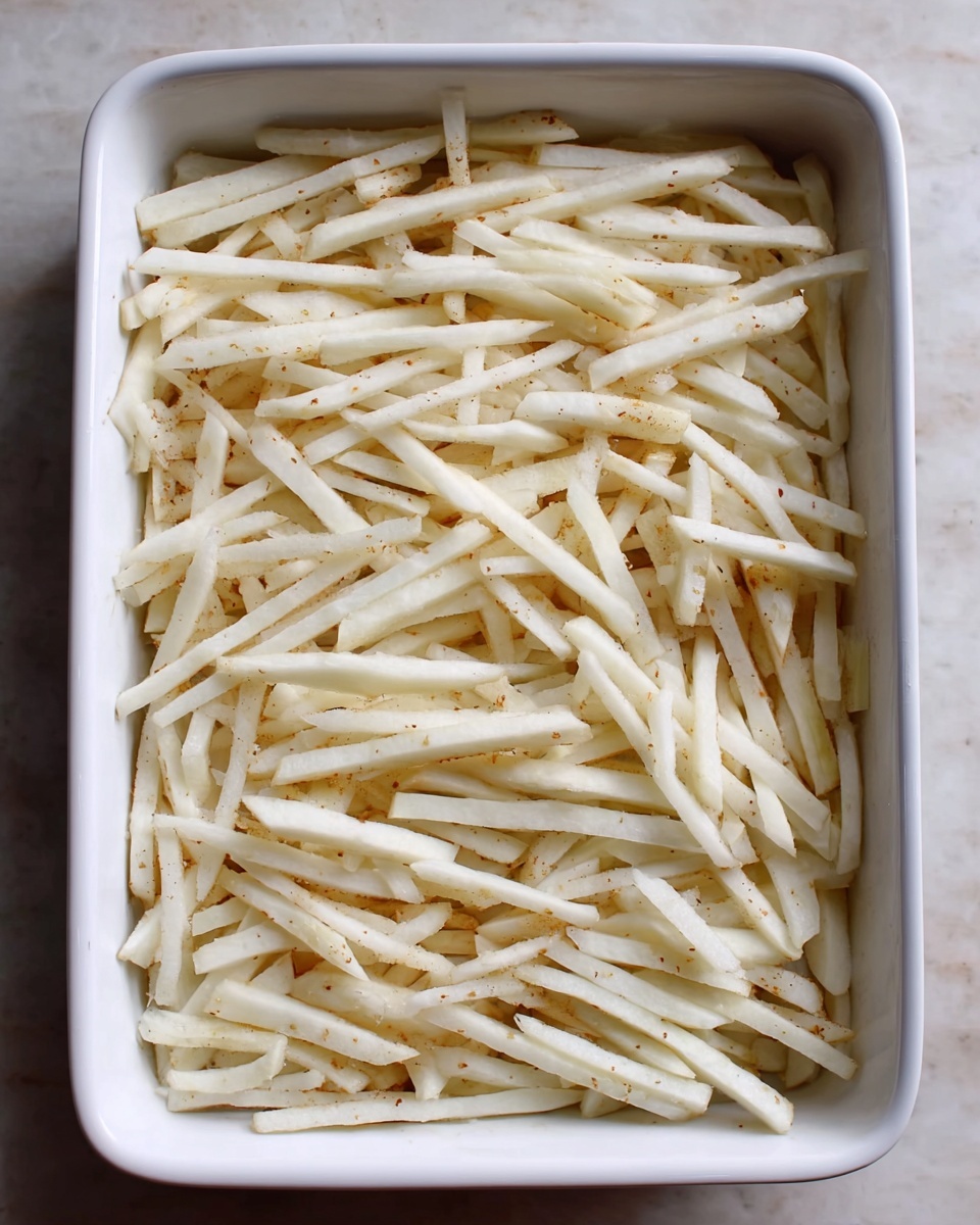 The image shows one layer of uncooked thin white potato sticks spread evenly in a white rectangular baking dish. The potato sticks are pale with slight beige spots, looking fresh and raw. The dish sits on a white marbled surface, and the potato sticks cover the entire dish without overlapping the edges. Photo taken with an iphone --ar 4:5 --v 7