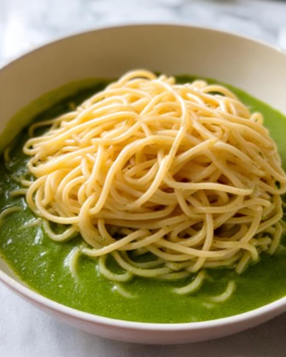 A close-up image shows a large white bowl filled with spaghetti pasta fully coated in thick, bright green pesto sauce. The sauce covers every strand of the pasta, giving it a creamy and slightly chunky texture from the herbs and nuts in the sauce. The spaghetti sits in a small mound at the center of the bowl, with the sauce spreading out evenly around the edges. The bowl rests on a white marbled surface. Photo taken with an iphone --ar 4:5 --v 7