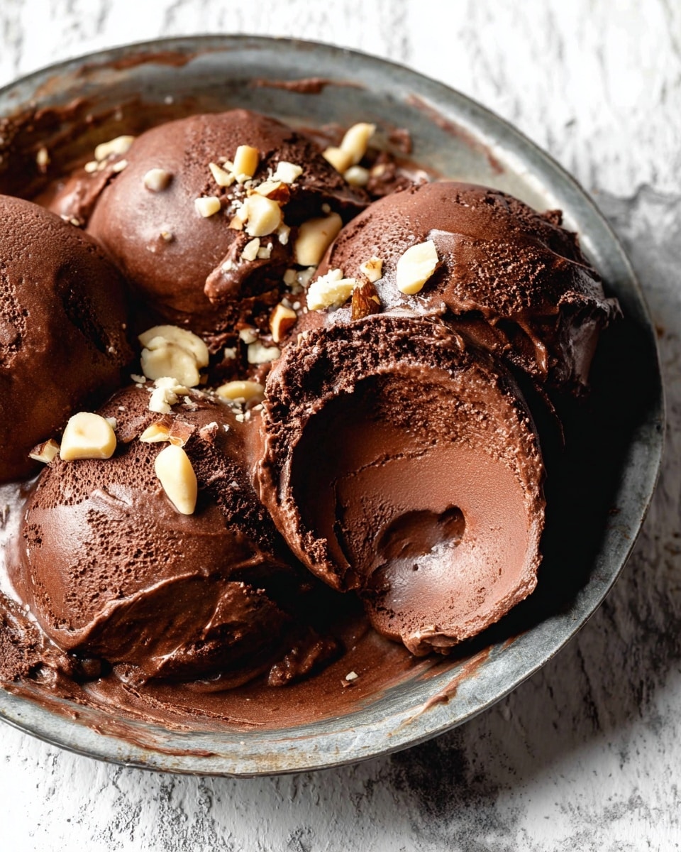 Two scoops of rich dark brown chocolate ice cream are placed side by side on a round white plate with a rough texture. The ice cream has a smooth, creamy surface with a slightly shiny and soft texture. Small pieces of chopped light beige nuts are sprinkled on top and around the ice cream, adding contrast. There are some thin dark chocolate shavings scattered lightly across the ice cream. The plate is set on a white marbled table surface. photo taken with an iphone --ar 4:5 --v 7