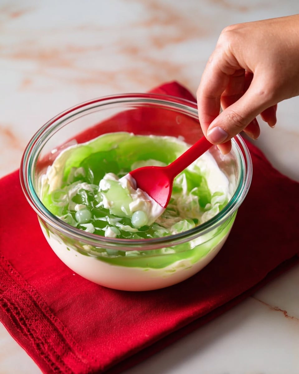 A white bowl sits on a white marbled surface with a red and white striped cloth underneath. Inside the bowl is a thick, pale green salad with a creamy, chunky texture, filling the bowl almost to the top. In the center, there is a dollop of white whipped topping, sprinkled with small pieces of chopped nuts that add a rough texture. Two bright red cherries with stems are placed on the whipped topping, one slightly tilted, adding a pop of color. In the background on the white marbled surface, a red bowl filled with chopped nuts and a small white bowl holding more cherries are visible, along with a golden spoon resting on the cloth. Photo taken with an iphone --ar 4:5 --v 7
