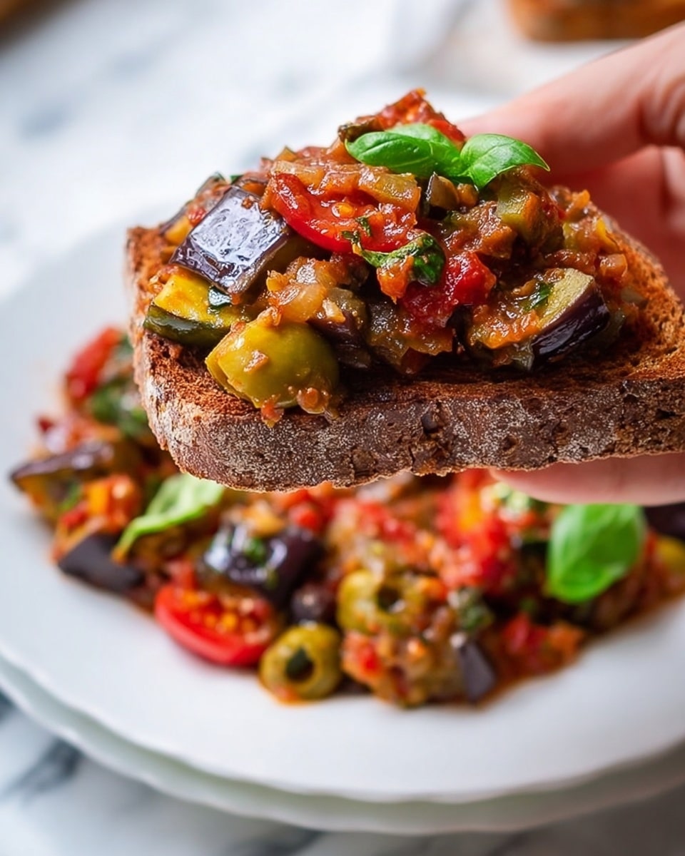 A close-up shows a woman's hand holding a slice of toasted brown bread topped with a colorful vegetable mix. The toast's layer is rough with a dark brown crisp edge. On top, there is a chunky vegetable mix with layers of shiny, cooked eggplant pieces in dark purple, soft red tomatoes, green olives, and bright green basil leaves. The background shows more of the vegetable mix spread out on a white plate, set against a white marbled surface. The colors are rich with a mix of reds, greens, and purples, showing a fresh, hearty dish. Photo taken with an iphone --ar 4:5 --v 7