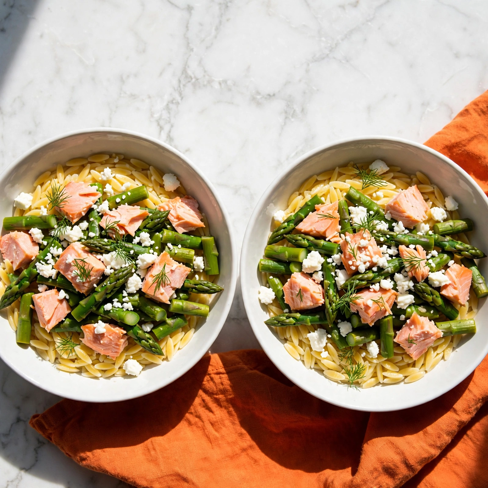 A white bowl filled with three main layers: the base layer is pale yellow orzo pasta, scattered throughout with light green roasted asparagus pieces on top, some showing slight charring. Layered above the asparagus are chunks of light pink cooked salmon, giving a soft, flaky texture. The final layer features small white dollops of creamy cheese, crumbled unevenly across the dish, with a few sprigs of fresh green dill sprinkled over everything. A metal fork rests on the right side of the bowl, partly inside. The bowl sits on a white marbled surface with a light orange cloth partially visible underneath the upper bowl. Photo taken with an iphone --ar 4:5 --v 7