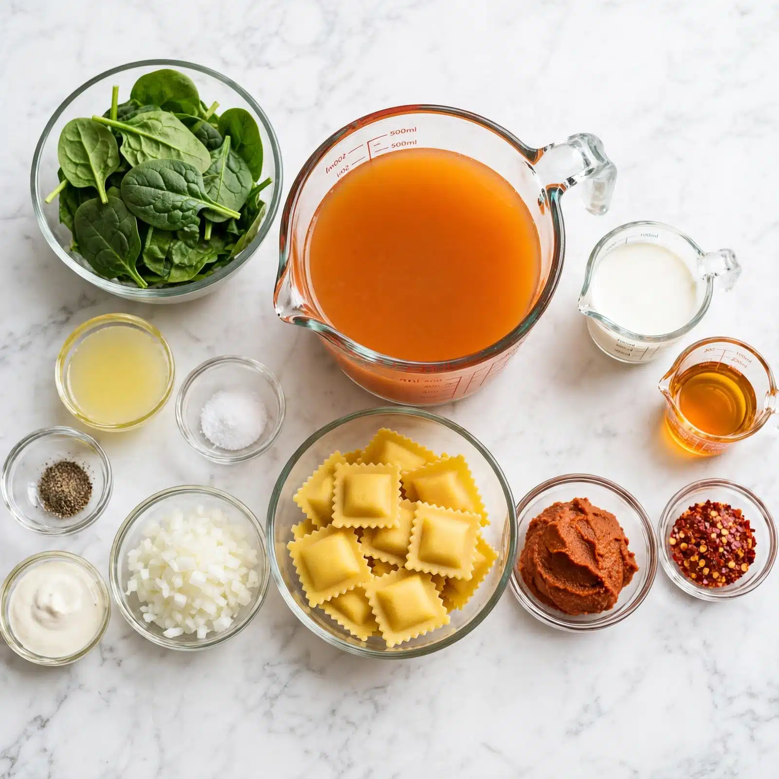 Creamy Sun-Dried Tomato & Spinach Soup with Ravioli Recipe 4 The image shows several small clear glass bowls and measuring cups arranged neatly on a white marbled surface, each containing different cooking ingredients. There is a medium bowl filled with fresh green spinach leaves on the top left, next to a large measuring cup filled with orange broth in the center. Below the broth, there is a medium bowl with uncooked ravioli pasta that is yellow with ruffled edges. To the right of the ravioli, there is a small bowl containing a reddish-brown paste, and next to it is a small bowl with red chili flakes. Moving left from the ravioli, there is a bowl of finely chopped white onions, a tiny bowl of salt, a small bowl of lemon juice, a small bowl with ground black pepper, and a tiny bowl with a creamy white sauce. Above these bowls on the right side, there is a smaller measuring cup with white cream and a tiny measuring cup with orange oil. The arrangement is clean and all bowls and cups are clear glass on the white marbled surface. Photo taken with an iphone --ar 4:5 --v 7