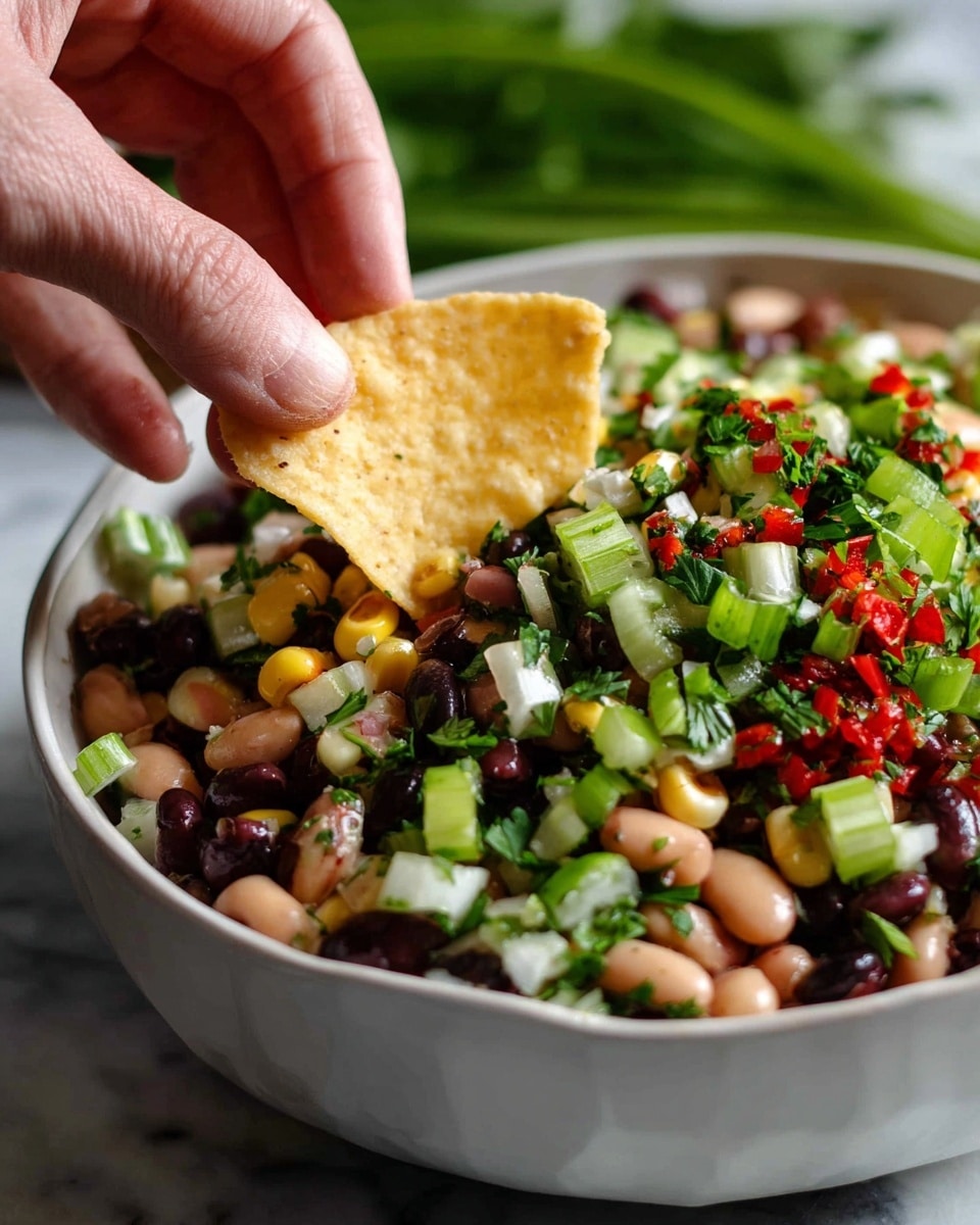 A white bowl sits on a white marbled surface filled with a colorful layered salad made of chopped green onions creating a bright green layer, black-eyed peas and black beans forming dark and light brown patches, small white corn kernels scattered throughout, diced red peppers adding bright red spots, and finely chopped green herbs mixed in for freshness. A silver spoon with worn, intricate designs rests in the bowl, slightly sunk into the salad. To the left, a bunch of fresh cilantro with green leaves peeks into the frame, while to the right, white tortilla chips are partly shown. In the bottom right corner, a glass contains lime wedges on the white marbled surface, with a soft cream cloth lying near the bowl. photo taken with an iphone --ar 4:5 --v 7