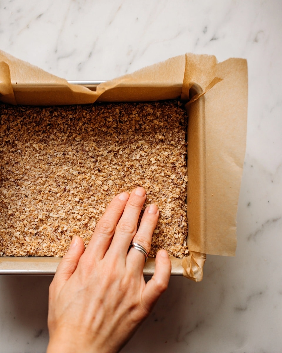 A close-up view of a rectangular baking pan lined with brown parchment paper folded around the edges. Inside the pan is a single thick layer of crumbly, light brown mixture with visible small bits of oats or nuts spread evenly and pressed down flat. A woman's hand with a silver ring on the ring finger is gently pressing the mixture into the pan from the bottom left corner. The pan is placed on a white marbled surface. photo taken with an iphone --ar 4:5 --v 7