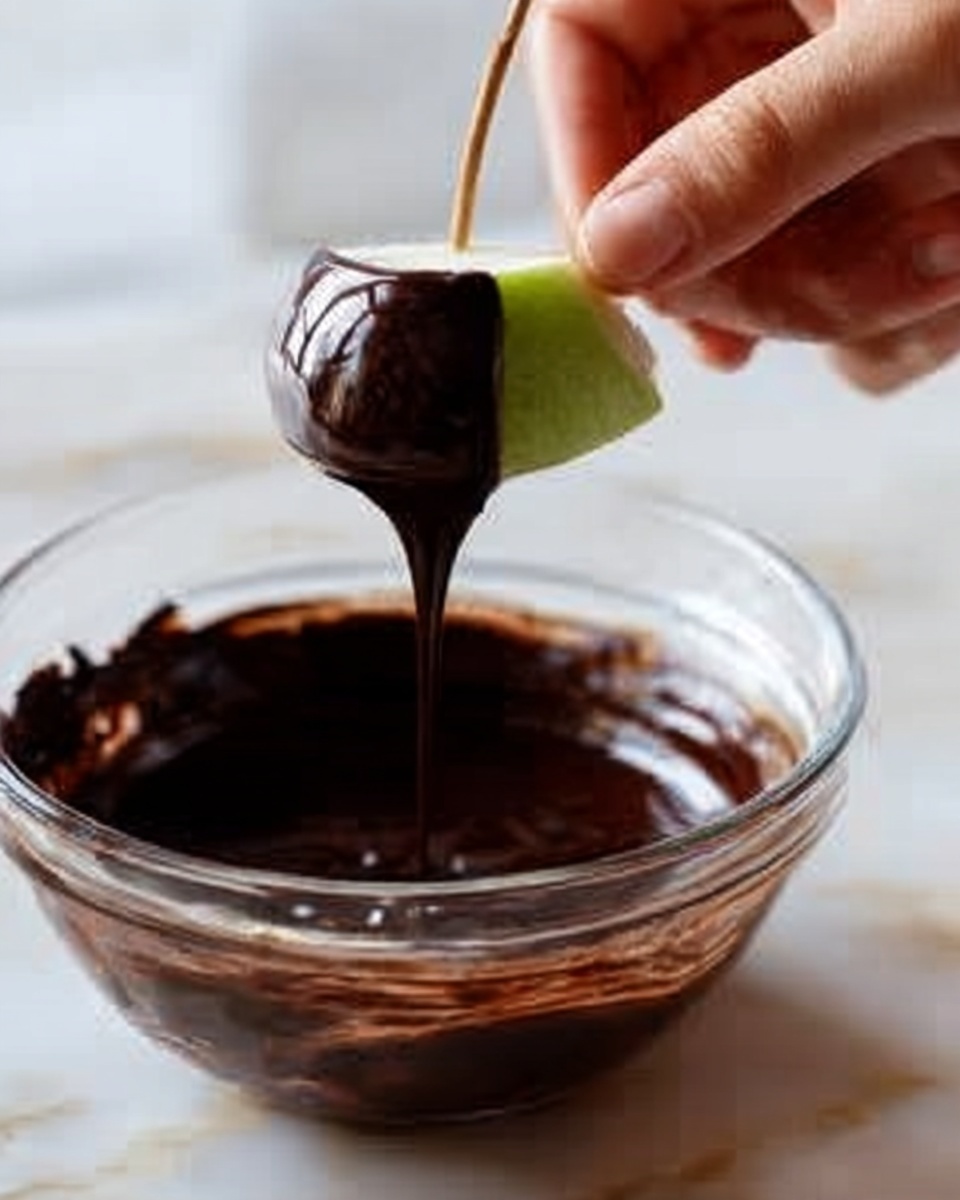 The image shows small treats with three clear layers. The bottom layer is a golden-brown pretzel base, flat and crunchy. Above it is a thick, dark chocolate layer that covers the sides and bottom half of an apple slice, with crushed nuts sticking to the chocolate. The top layer is a bright green apple slice with smooth skin. Each treat is held together by a small wooden stick placed vertically through the center. The treats are placed on a round white plate, all set against a white marbled surface. The lighting is soft and natural, highlighting the texture of the chocolate and nuts. photo taken with an iphone --ar 4:5 --v 7
