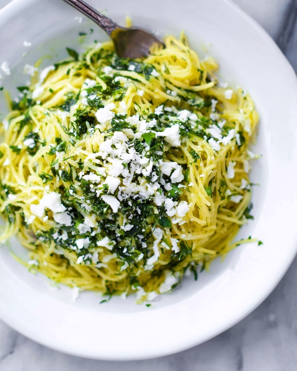 A close-up view of a single white plate with a rounded heap of thin, spiral-shaped yellow noodles mixed with bright green chopped herbs spread evenly across them. Small white soft cheese crumbles sit on top and around the edges of the noodles. The plate rests on a surface with a white marbled texture, and fresh green herbs and a lemon are visible blurred in the background. photo taken with an iphone --ar 4:5 --v 7