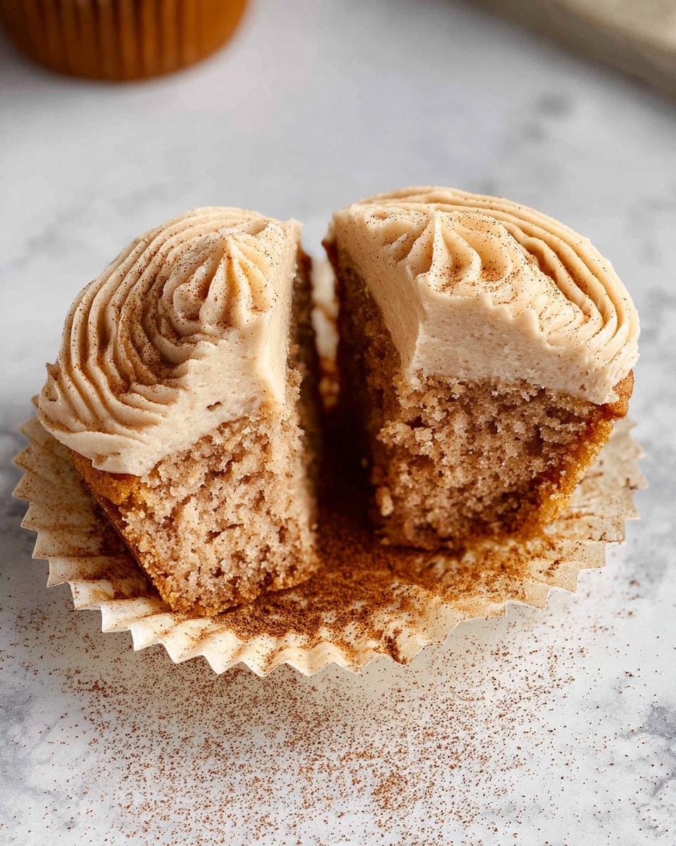 A single cupcake with one visible layer of light brown cake inside a ridged, white paper wrapper sits on top of a dark gray fluted metal base, all set on a white marbled surface. The cupcake has a thick swirl of creamy beige frosting on top, dusted lightly with fine brown specks, giving it a soft and smooth texture. A blurred glass of milk and cinnamon sticks are in the background, softly lit with a neutral gray backdrop. photo taken with an iphone --ar 4:5 --v 7