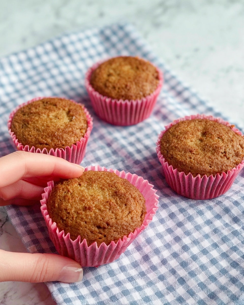 Four brown muffins with a rough, baked texture are placed on a blue and white checkered cloth over a white marbled surface. Each muffin sits in a ruffled pink paper cup. One muffin is being held by a woman's hand from the bottom left, showing the light skin tone. The muffins have a slightly cracked top and a warm, golden-brown color. photo taken with an iphone --ar 4:5 --v 7