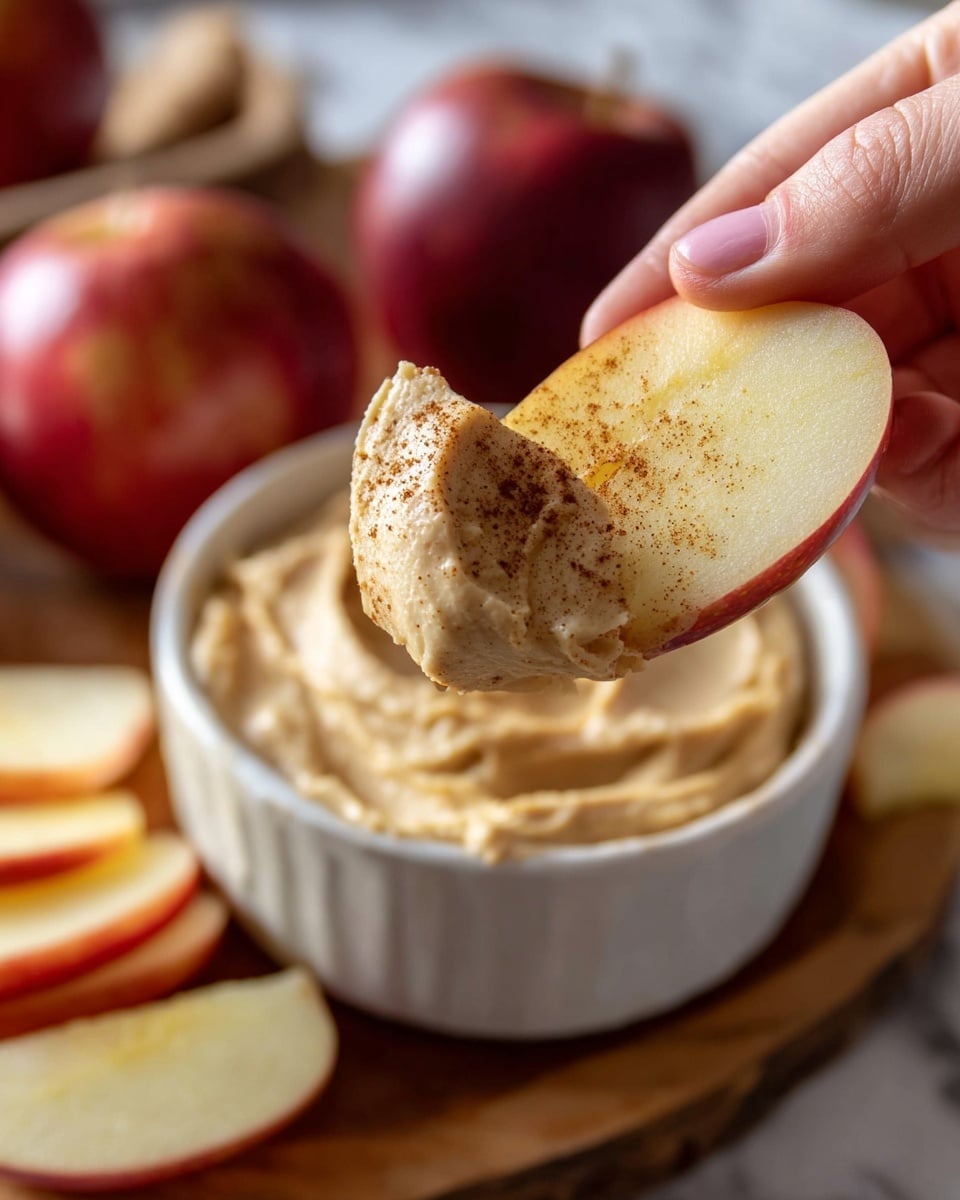 A white ramekin filled with a creamy light brown dip that is swirled smoothly on top and sprinkled with a light dusting of cinnamon or cocoa powder. The ramekin sits on a wooden cutting board surrounded by several red apples, some whole and some sliced into wedges revealing their pale yellow insides. The surface beneath the cutting board is a white marbled texture. Photo taken with an iphone --ar 4:5 --v 7