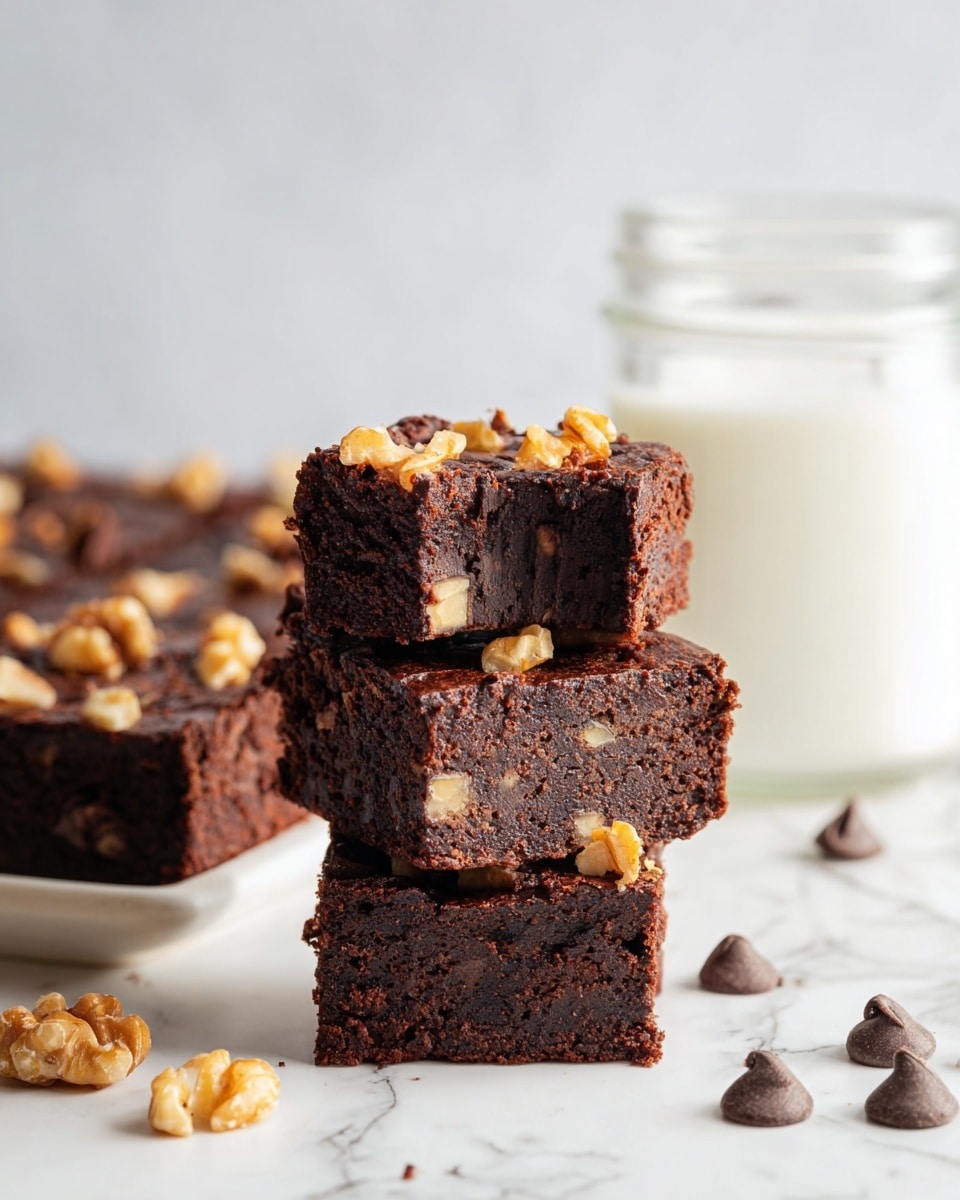 The image shows a stack of three dark brown brownies with a dense and moist texture, each topped with small pieces of light golden walnuts. The middle brownie on the top layer has a bite taken out, revealing a rich, chocolatey inside with visible chunks. In the background, there is a glass jar filled with white milk and a blurred white mug, both sitting on a white marbled surface. There are a few chocolate chips scattered around the brownies in the foreground. photo taken with an iphone --ar 4:5 --v 7