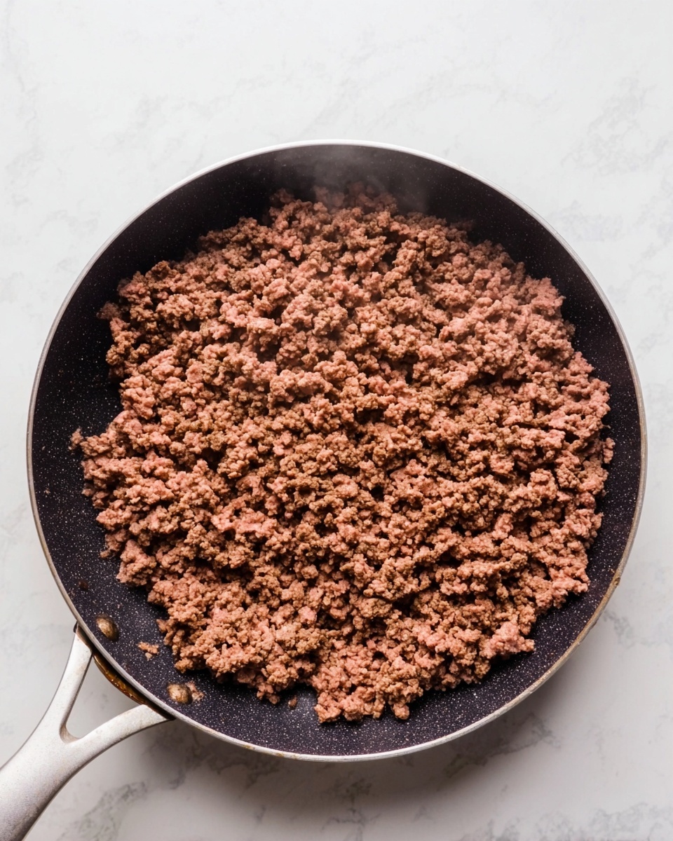 A black pan with a white handle filled with cooked ground beef with small crumbly pieces, steaming lightly. The pan sits on a white marbled surface, and the cooked beef is spread evenly across the pan, showing a rich brown color with a slightly coarse texture. photo taken with an iphone --ar 4:5 --v 7