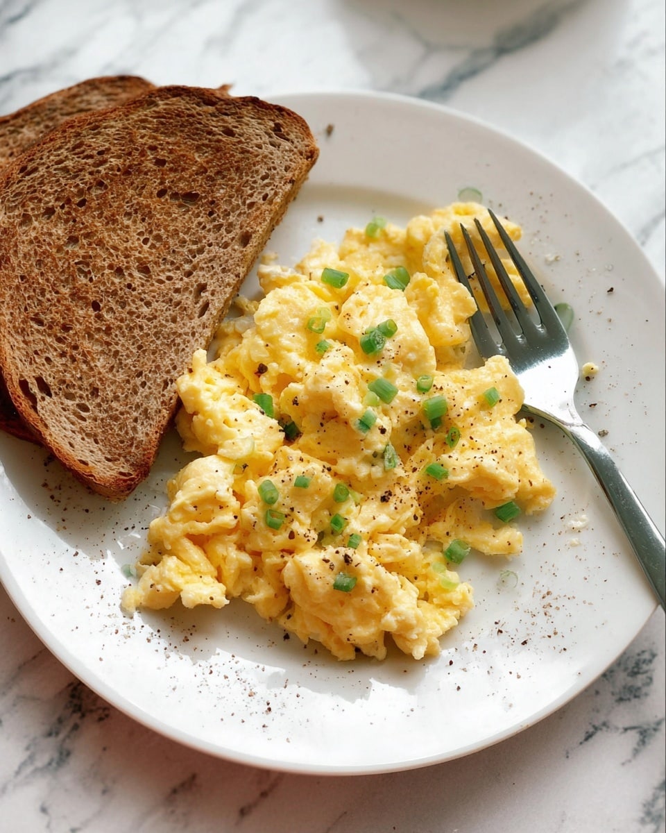 A white plate with a thin brown edge holds a simple breakfast of two main layers: on the left, a single slice of toasted bread, golden-brown and evenly toasted; on the right, a soft pile of scrambled eggs with a creamy yellow color, lightly sprinkled with small green onion pieces and black pepper flakes. A silver fork rests on the plate near the eggs. The setting includes a white marbled surface with a small glass bowl of white cottage cheese in the foreground and an egg and spice jars blurred in the background. photo taken with an iphone --ar 4:5 --v 7