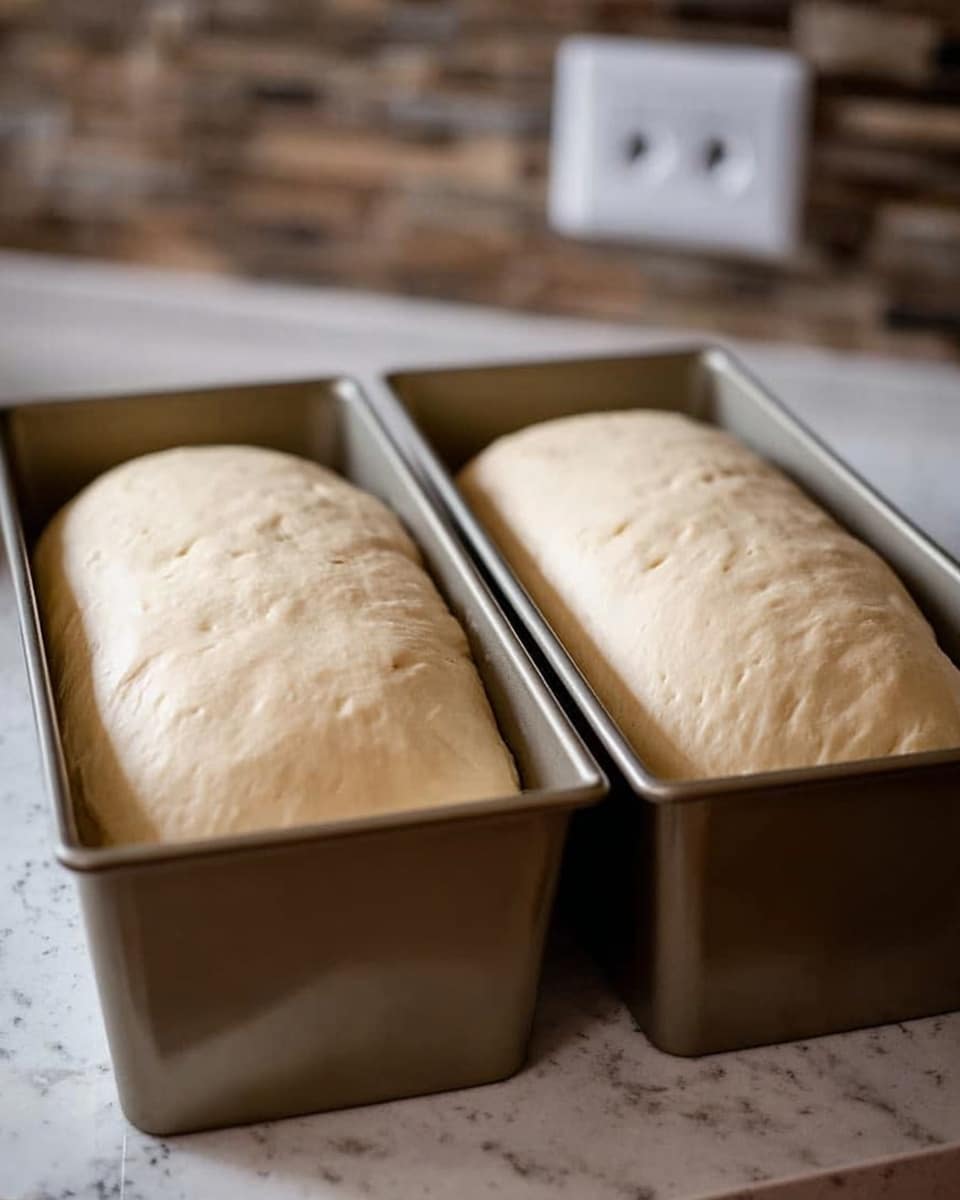 The image shows two golden metal loaf pans side by side on a white marbled surface, each filled with light beige dough that has puffed up well above the edges of the pans. The dough has a smooth texture with small dimples and a slightly shiny surface, indicating it has risen fully. The background is blurred, featuring a stone wall and a power outlet. The focus is on the soft, swollen dough in the pans, ready to be baked. Photo taken with an iphone --ar 4:5 --v 7