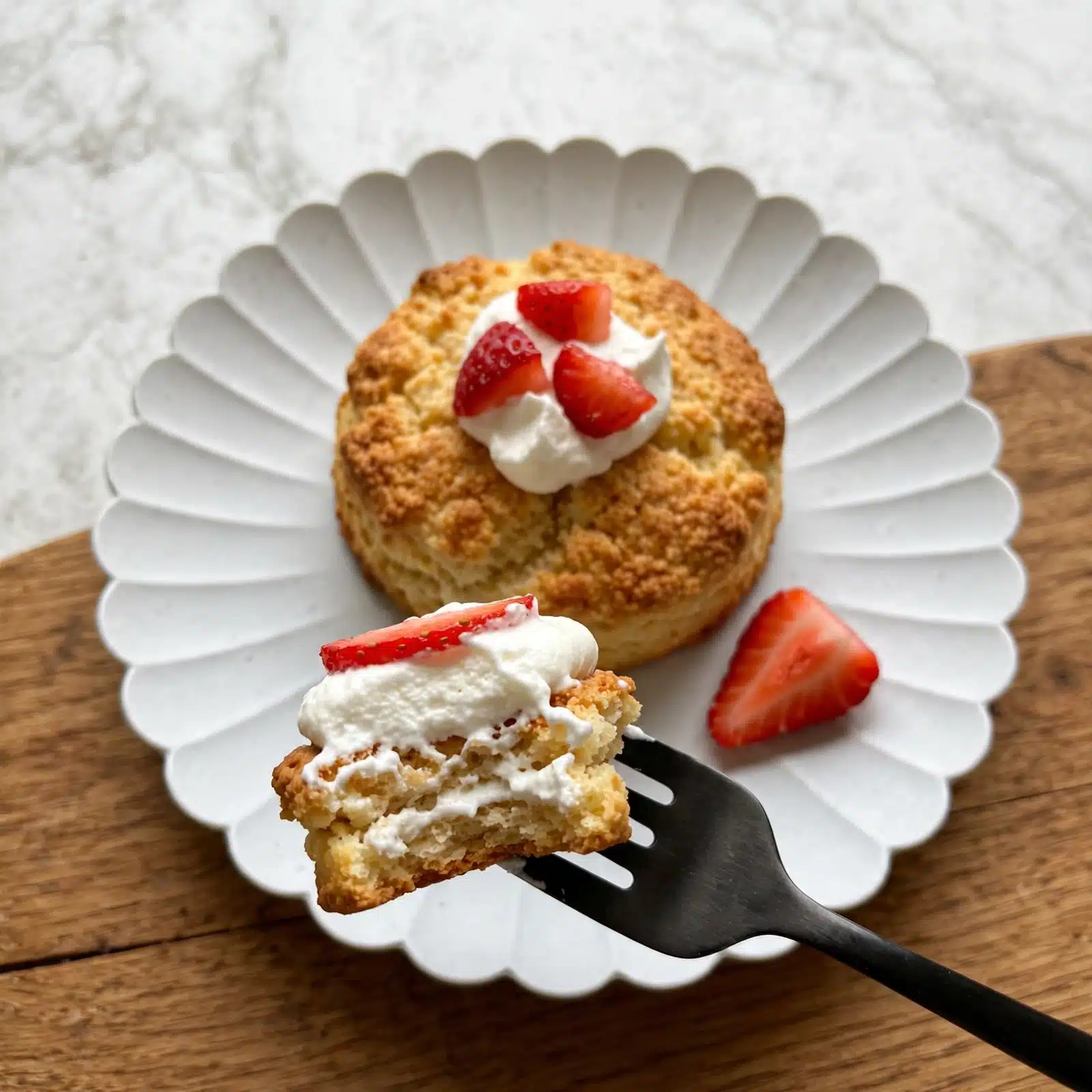 Classic Strawberry Shortcake Recipe 5 A shortcake with two thick layers of golden, crumbly biscuit sits on a white scalloped plate. Between the biscuit layers, there are fresh bright red sliced strawberries and a layer of smooth white cream. More strawberries and a dollop of cream sit on top of the biscuit. The plate is on a wooden surface with a white marbled background. Photo taken with an iphone --ar 4:5 --v 7