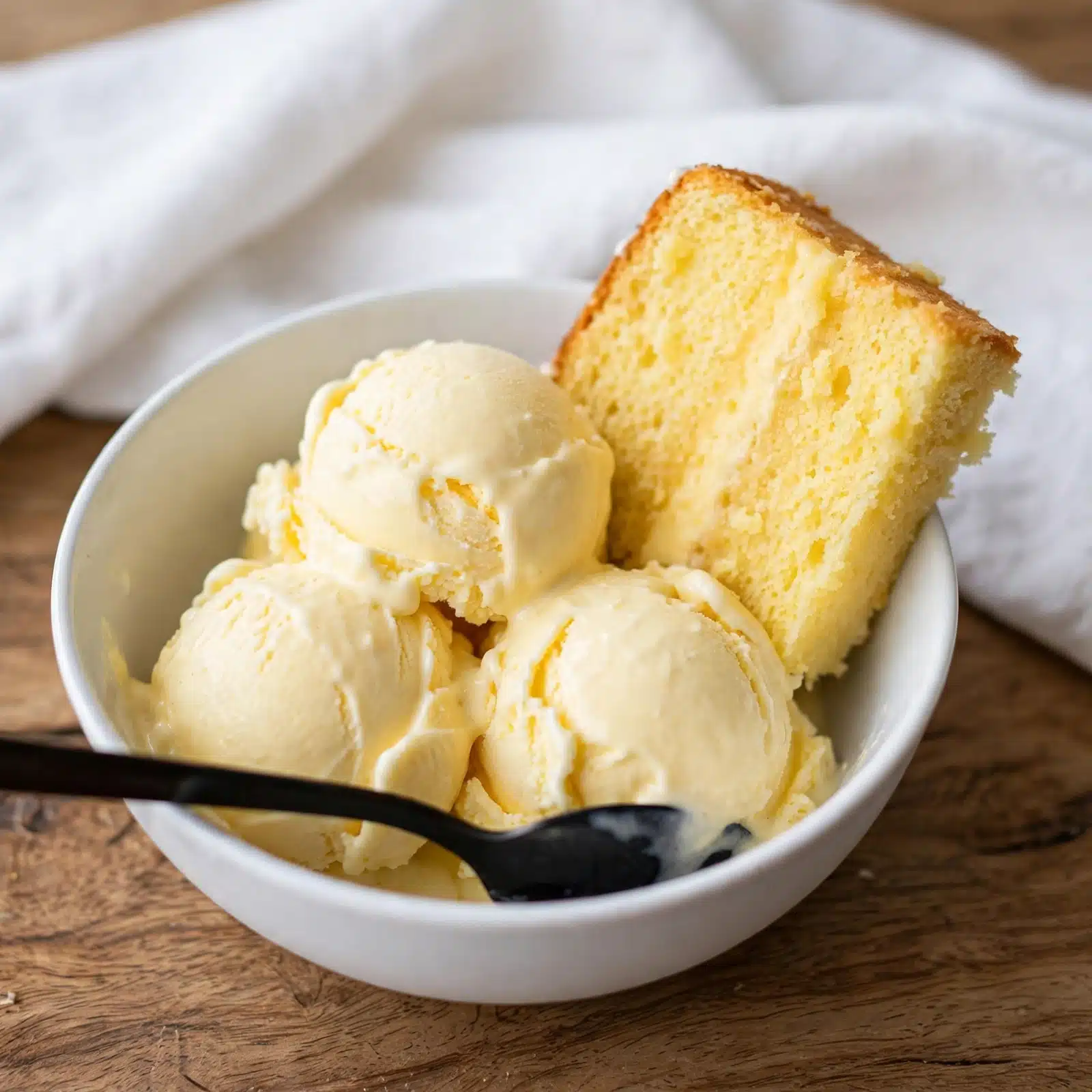 Homemade Cornbread Ice Cream Recipe 5 A white bowl filled with three scoops of pale yellow ice cream sits on a wooden table with a white cloth in the background. There is a thick slice of yellow cake leaning against the ice cream inside the bowl. A black spoon rests in the bowl, partially covered by the ice cream. Photo taken with an iphone --ar 4:5 --v 7