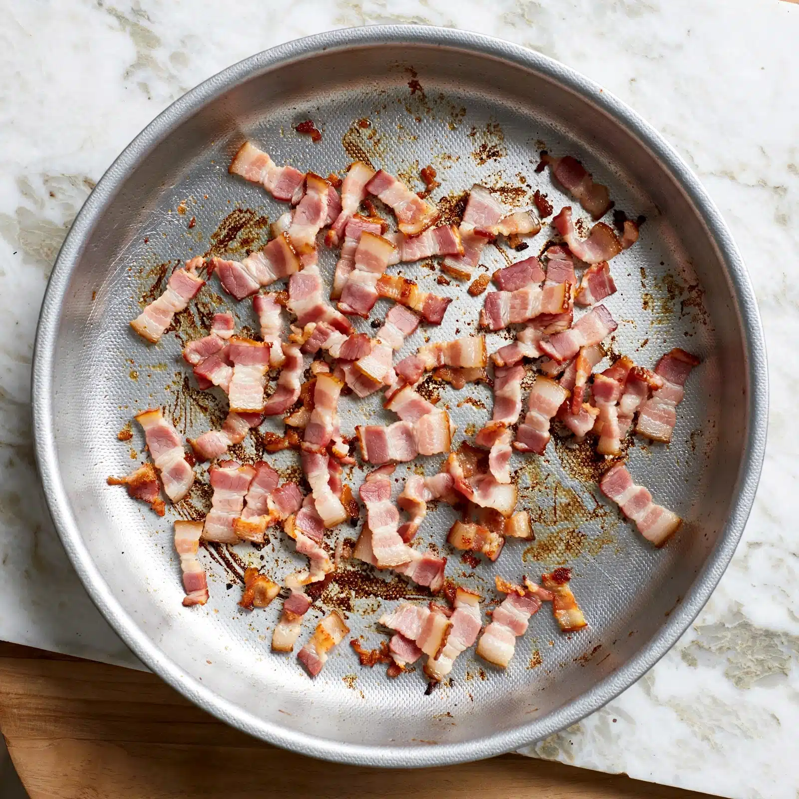 A round silver pan holds small strips of cooked bacon scattered unevenly across its surface. The bacon pieces are a mix of light pink and slightly browned edges, laying on a textured silver pan bottom with brown cooked marks. The pan rests on a white marbled textured surface with a light brown wooden board partially visible underneath. In the upper left corner, a bright green circle with the black number 