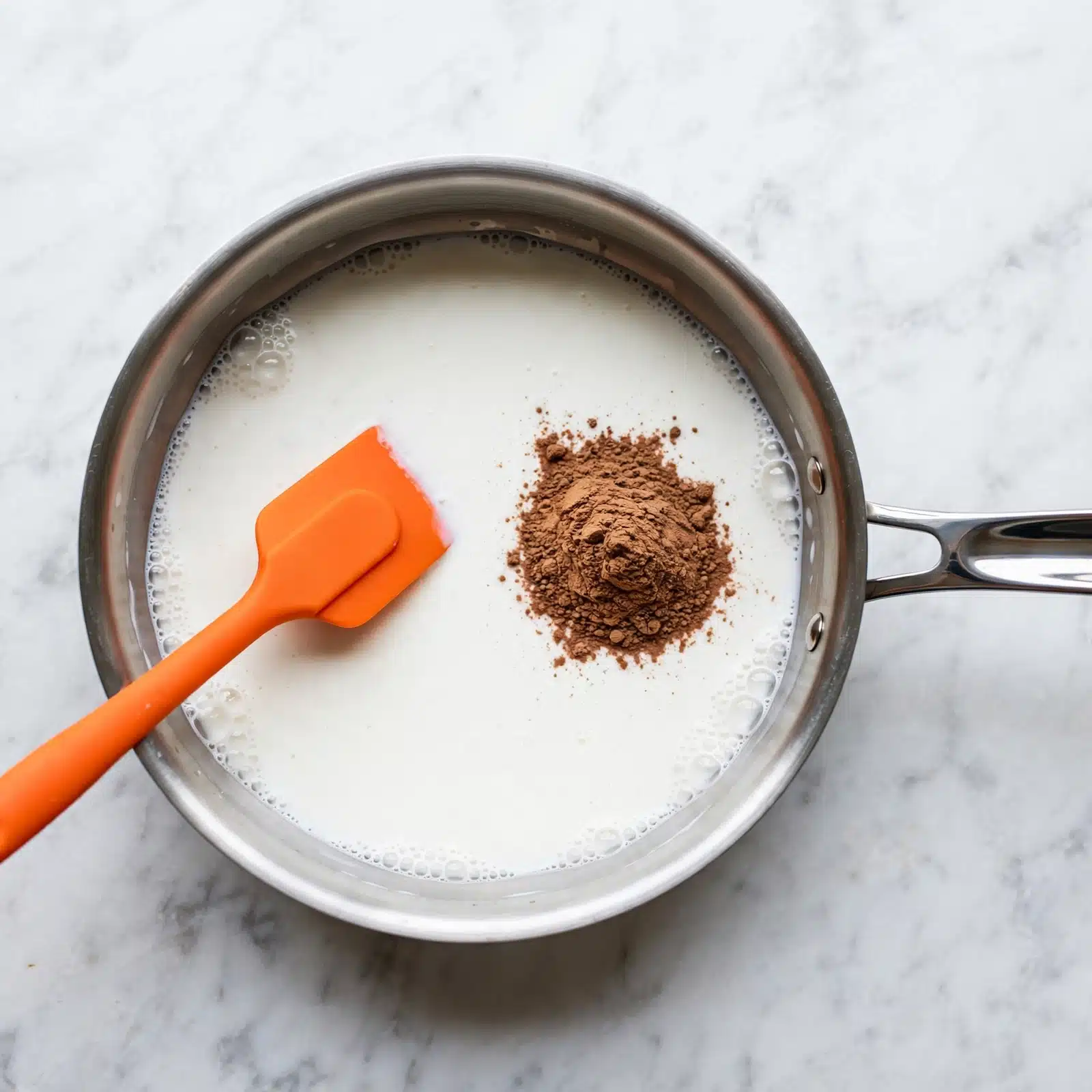 Limbers de Coco: Creamy Puerto Rican Coconut Ice Pops Recipe 4 A round silver pan with a shiny handle on the right sits on a white marbled surface. Inside the pan, there is a layer of white liquid with some bubbles near the edges. On top of this liquid, to the right of the center, there is a small pile of brown powder. A bright orange spatula lies inside the pan, partially resting on the liquid near the left side. The background is a smooth white marbled texture. Photo taken with an iphone --ar 4:5 --v 7